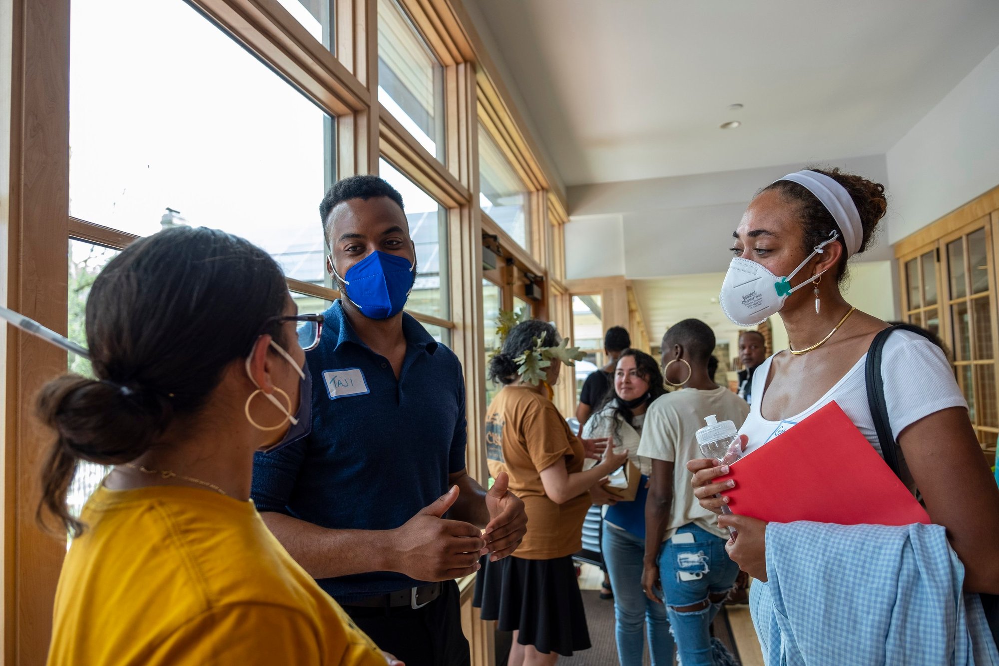 A group of people talk in a hallway. They are all wearing masks and appear to be making friendly conversation.