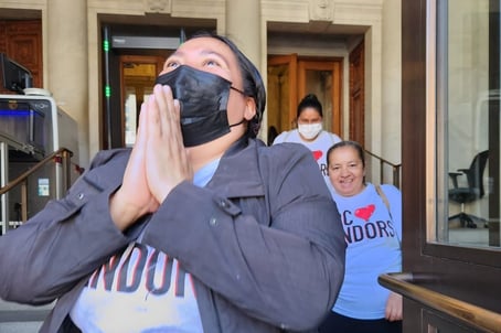 A woman looks joyous and has her hands clasped in prayer, looking up at the sky. She wears a mask and a shirt that says "DC loves vendors." In the background, a couple of other people look joyous with the same shirt on.