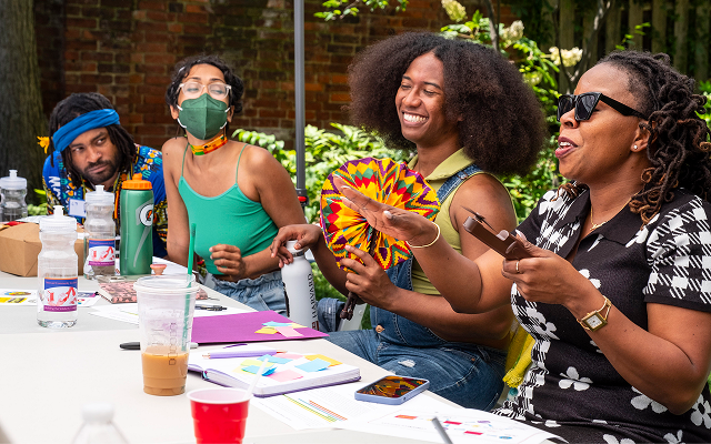 Four people sit next to each other at a table and look happy, they appear to be mid conversation. One holds a brightly colored fan, and all are dressed as though it is hot outside. One person wears a mask.