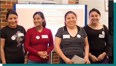 Four people stand in front of a screen showing a slide of text. The people wear name tags and look happily at the camera.