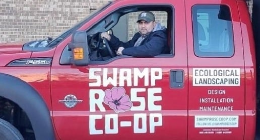 A worker owner sits in a new red truck with a large logo for Swamp Rose Co-op on the side. Another sign on the truck reads "Ecological Landscaping."