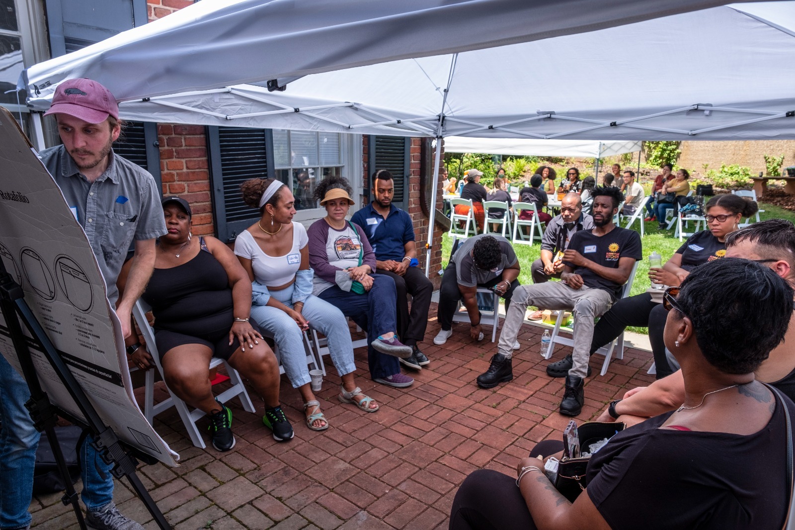 A diverse group of people sitting in a circle having a conversation outside under a tent. It's a sunny day. 
