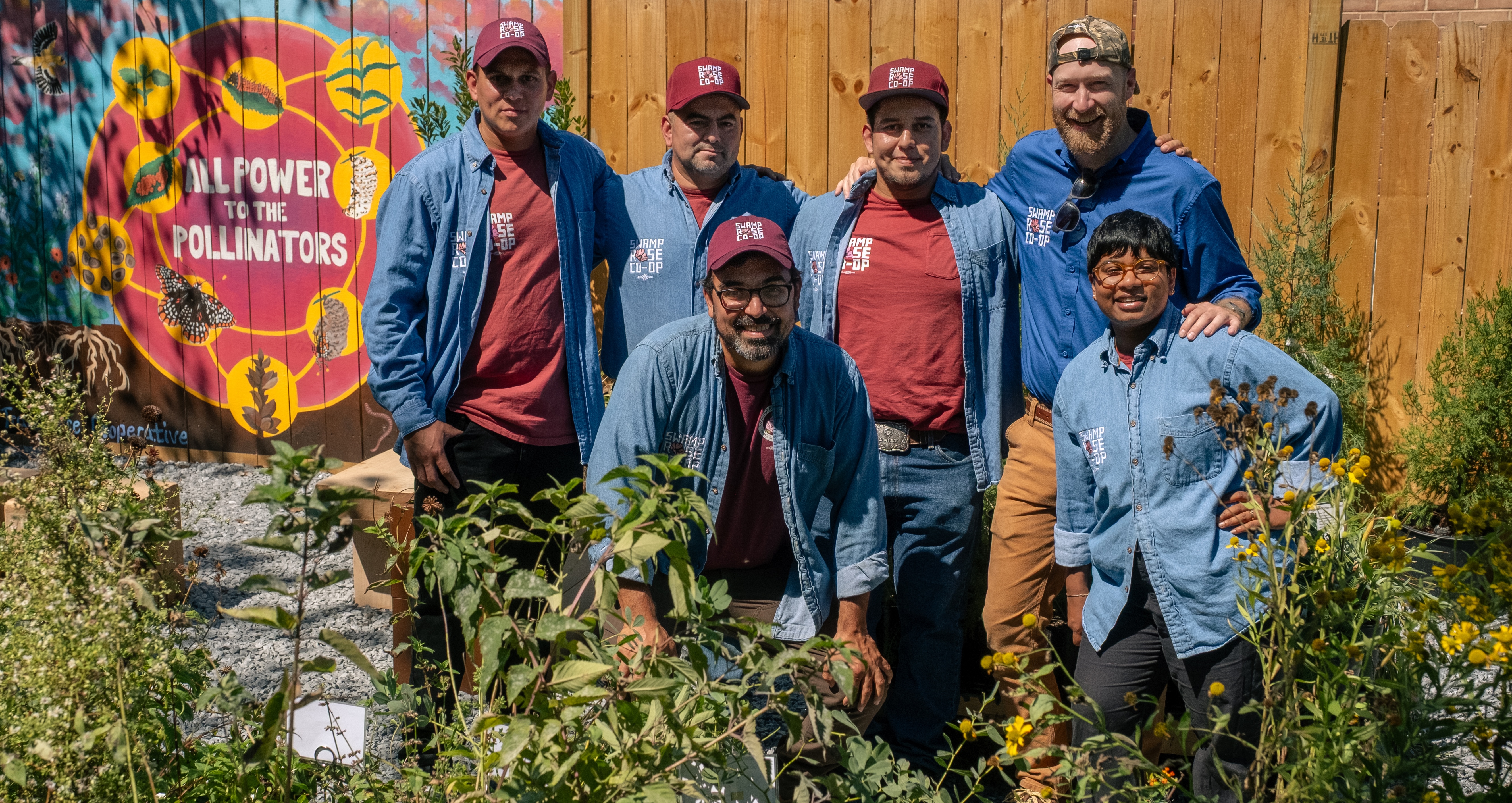 Swamp Rose Co-op members pose for a group photo wearing denim Swamp Rose button down shirts and maroon Samp Rose baseball caps. They're standing outside in a community garden on a sunny day. 