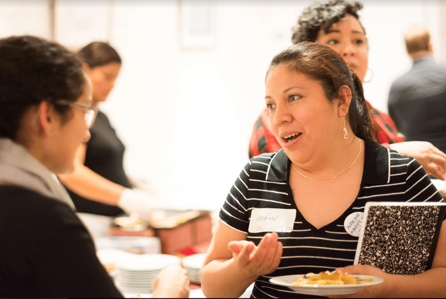 Two people appear in mid conversation in a room where other people are also present. The two having the conversation look engaged, with one person talking while holding a notebook and a plate of food, and the other listening.