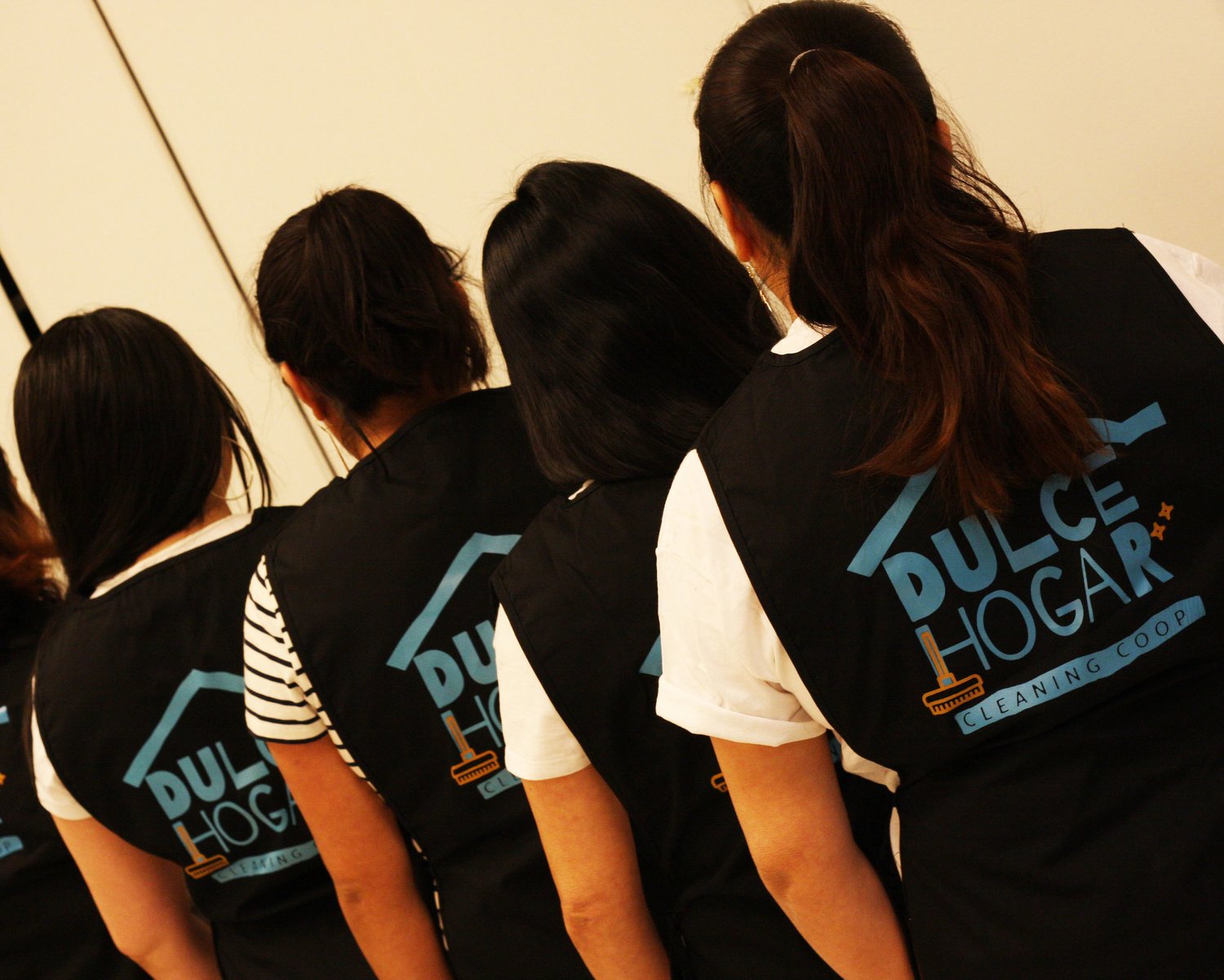 Four people stand with their backs facing the camera near a wall. They wear identical vests, which say "Dulce Hogar Cleaning Co-op" on the back.